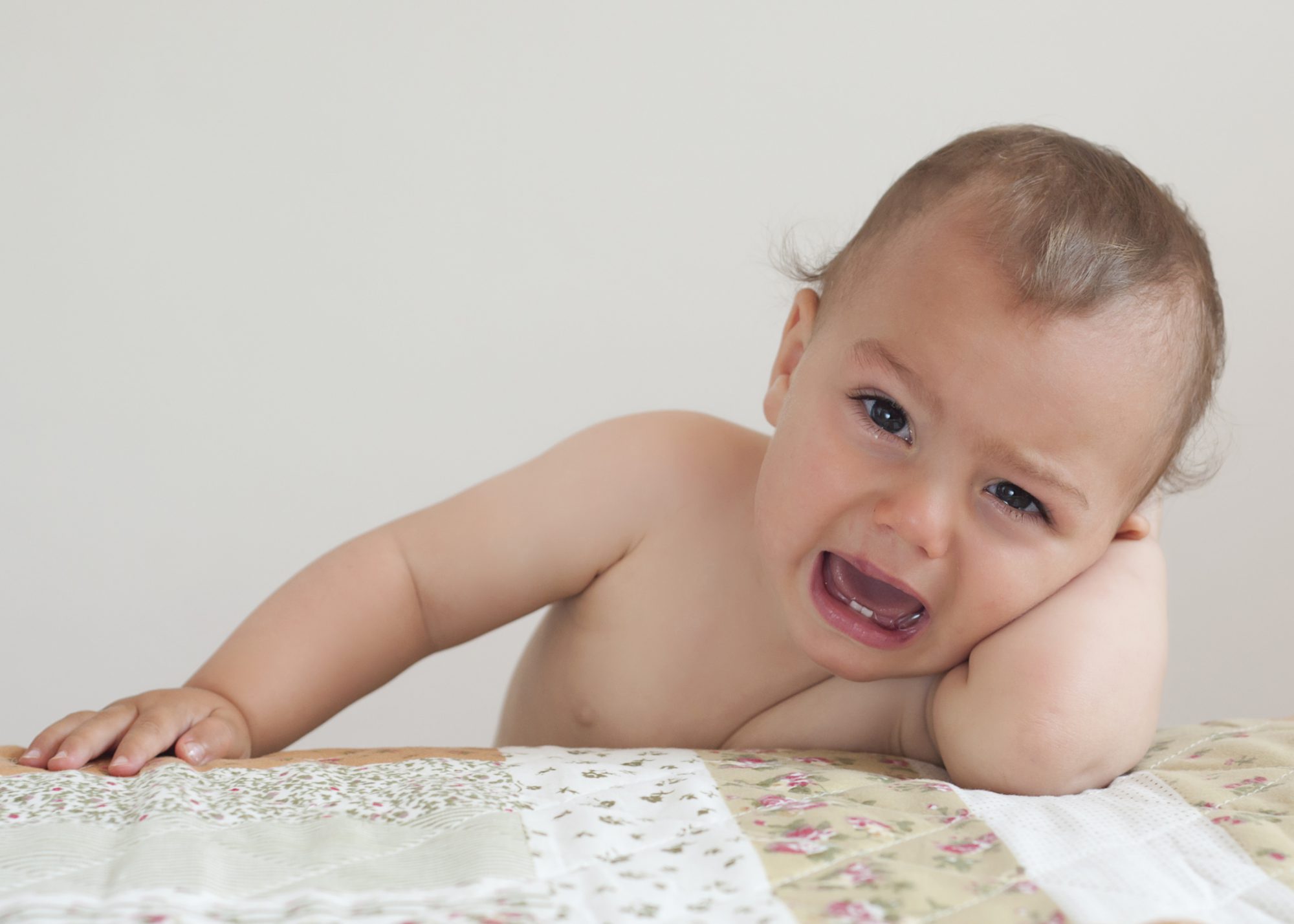 Crying baby or toddler child, boy or girl, at home leaning on bed.