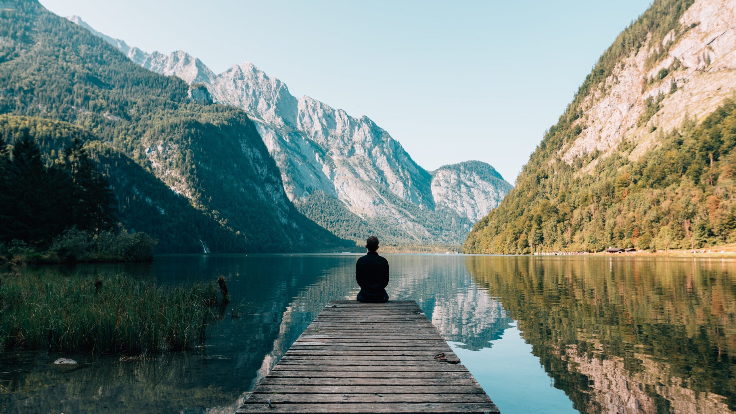 Contemplative person with their back to us sitting at the end of a pier regarding the mountains rising out of the far side of the lake.